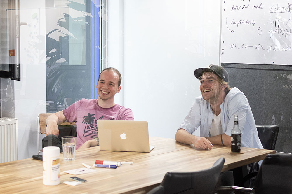 Two young male employees of shopping24 are sitting in a meeting room in front of their laptops and working on a presentation